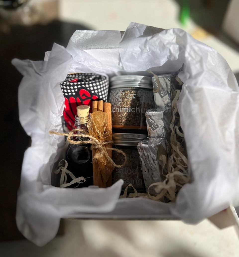 Gift basket with various items including a mini oil bottle, chocolate, various spices, tea towel and wrapped packages on white tissue paper.